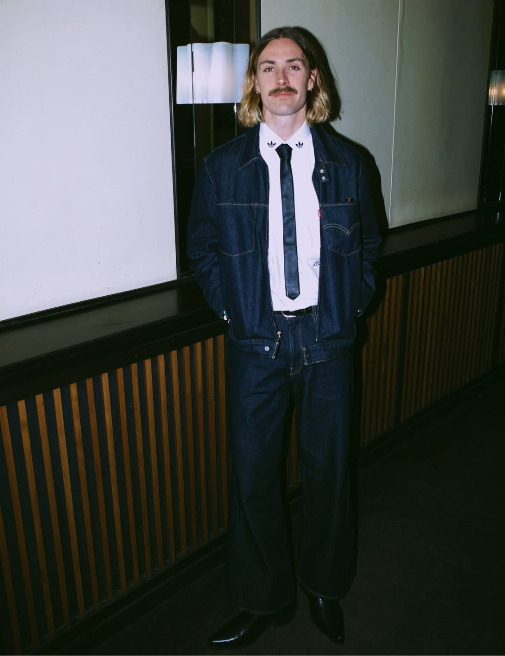 Jackson Irvine posa sereno com jaqueta e calça jeans amplas, camisa branca, gravata preta e botas em ambiente sóbrio.