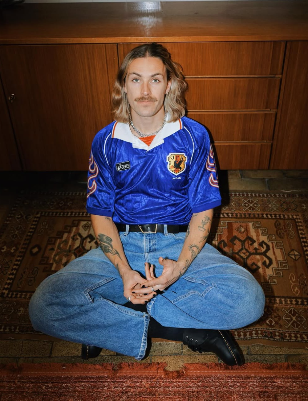 Jackson Irvine posa sentado no tapete com camisa azul brilhante, jeans amplos e botas pretas em clima retrô sereno.