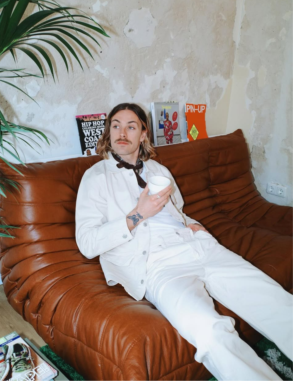 Jackson Irvine relaxa em sofá de couro caramelo com look branco amplo e lenço preto, clima vintage sereno.