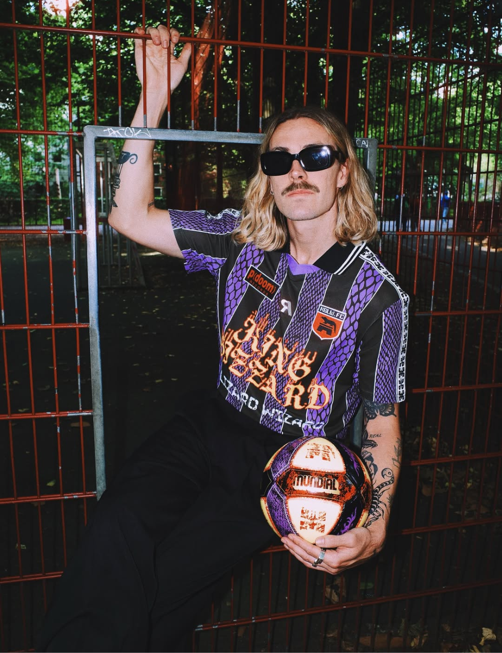 Jackson Irvine posa com camisa esportiva roxa e preta, óculos escuros e bola; grade vermelha cria clima urbano confiante.