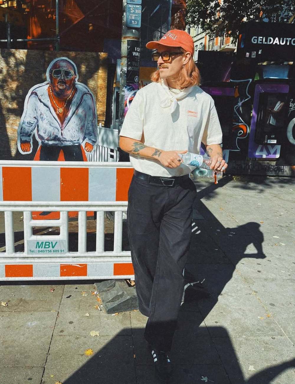 Jackson Irvine posa na rua com polo off-white, calça preta ampla e boné coral, em clima urbano descontraído.