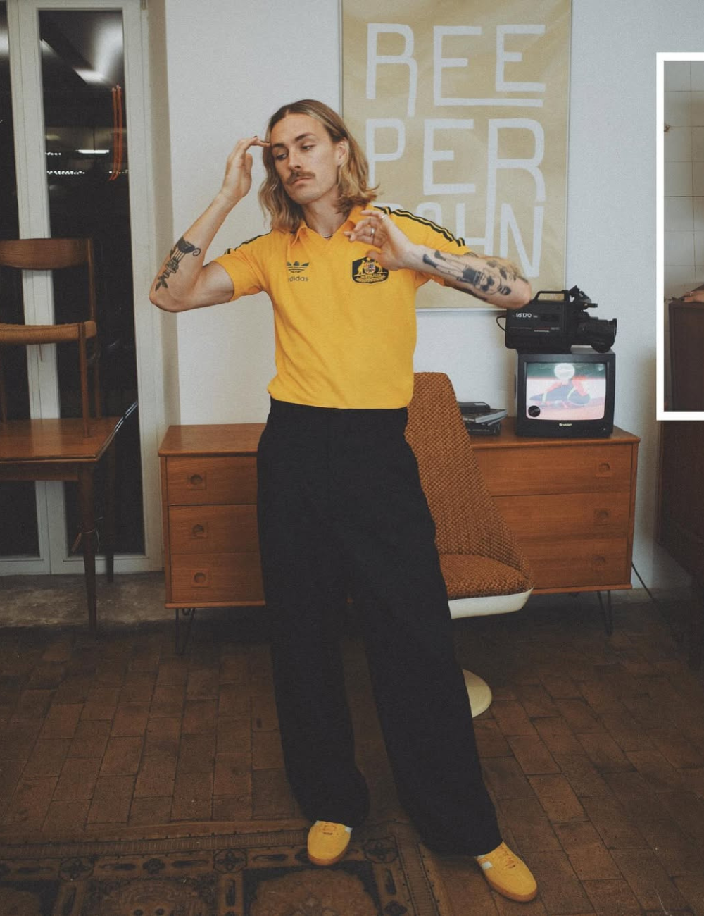 Jackson Irvine posa de polo amarelo retrô, calça preta ampla e tênis, em sala vintage de clima descontraído.