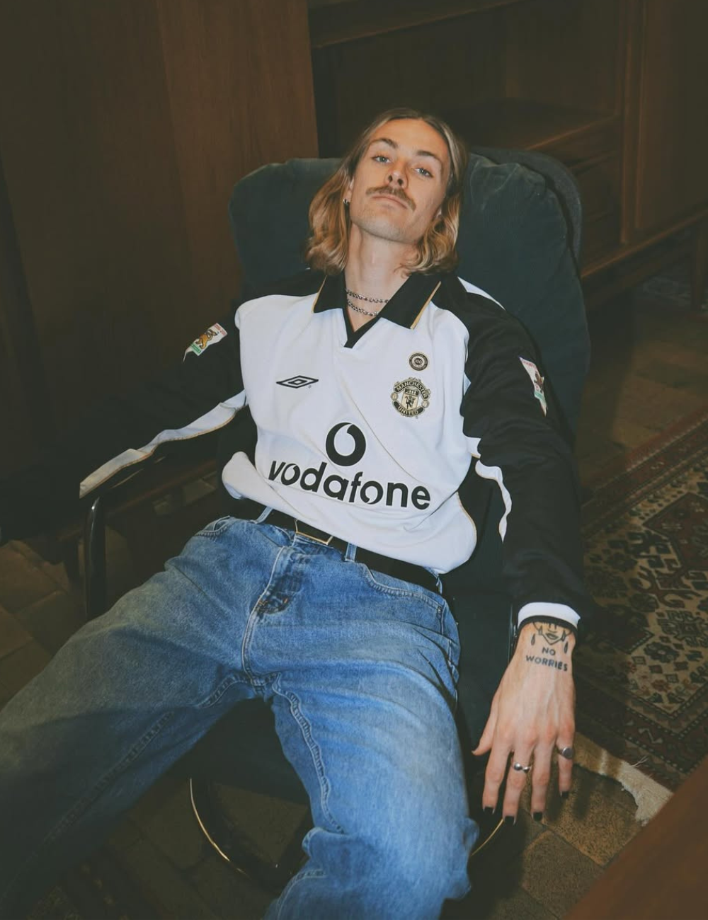 Jackson Irvine posa reclinado em camisa retrô do Manchester United, jeans largos e clima despojado.