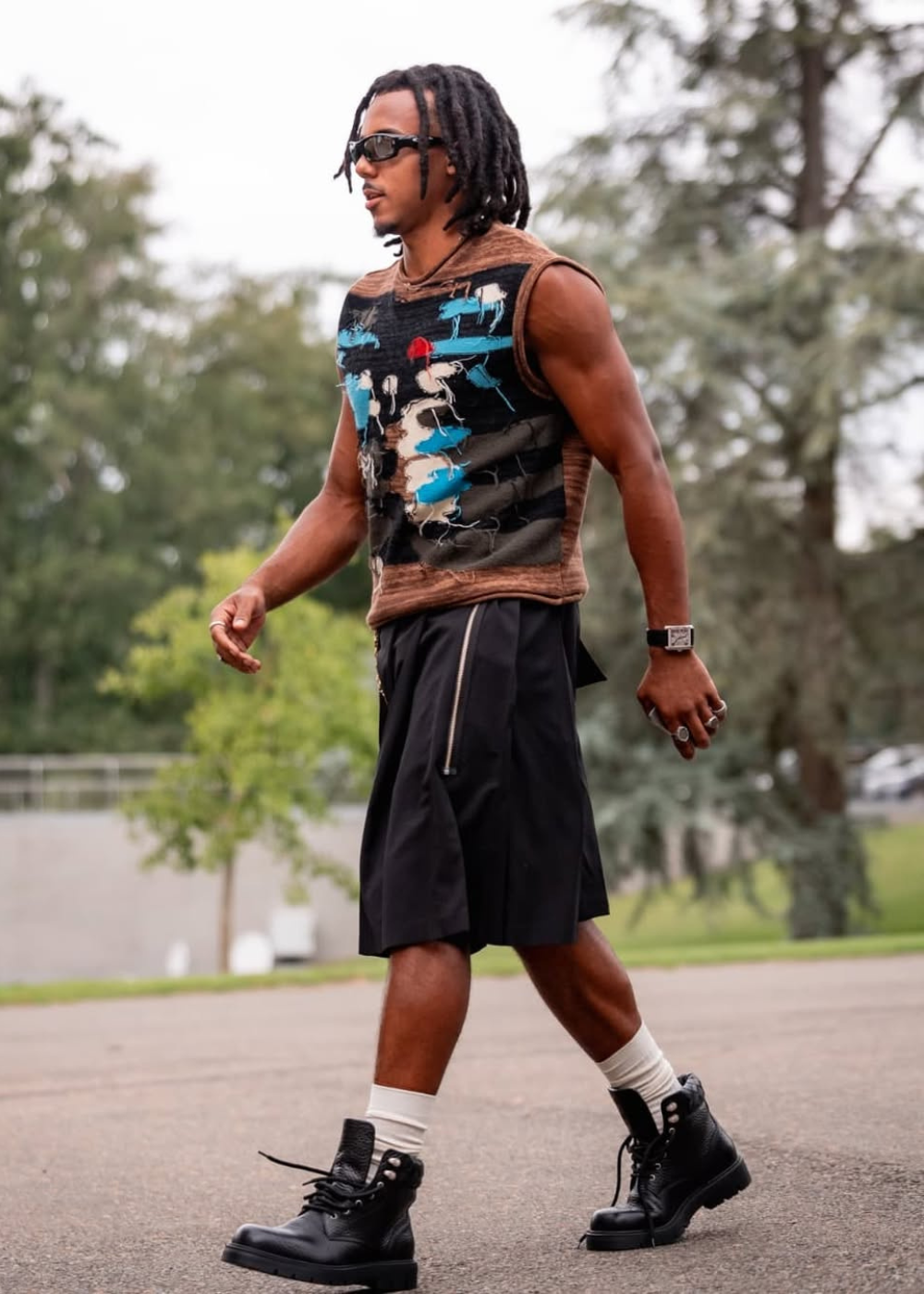 Jules Koundé caminha ao ar livre com regata estampada, bermuda ampla preta e coturnos, em visual moderno e confiante