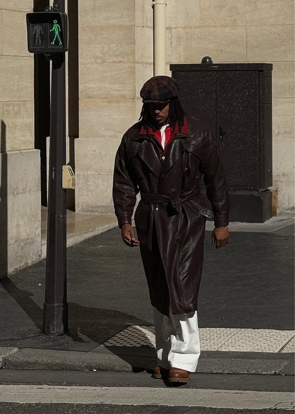 Jules Koundé cruza a rua com trench de couro marrom, boina xadrez e calça branca ampla em clima urbano sóbrio.