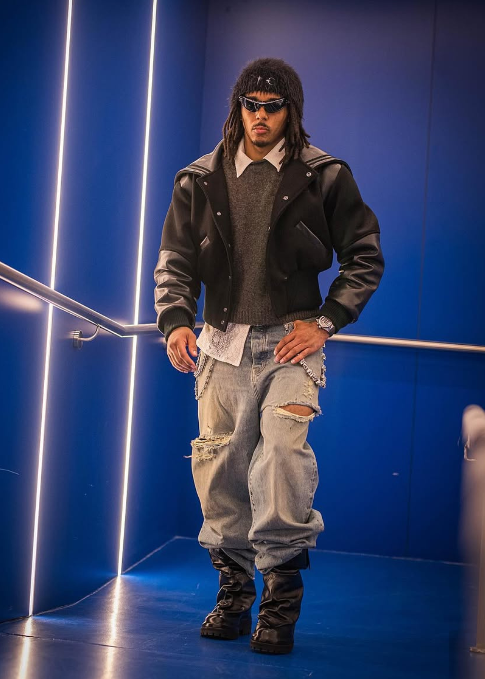 Jules Koundé posa com jaqueta preta estruturada, jeans rasgado amplo e óculos futuristas em cenário azul de clima ousado.