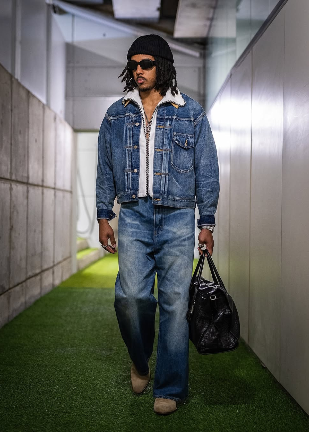 Jules Koundé caminha com jaqueta e calça jeans amplas, gorro preto, óculos escuros e bolsa preta em clima urbano confiante
