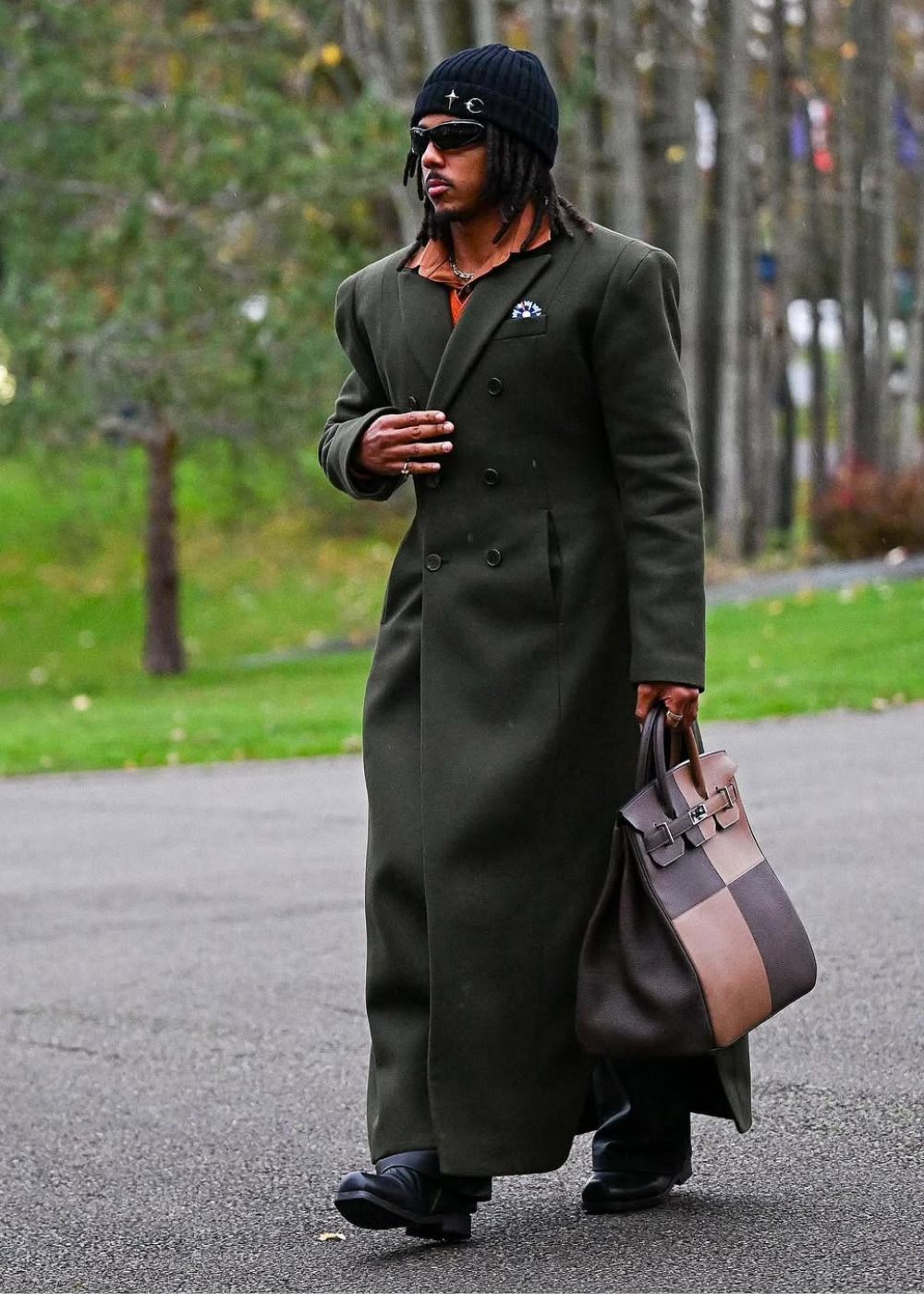 Jules Koundé caminha com sobretudo longo verde-oliva, gorro e óculos pretos, bolsa de couro marrom e elegância sóbria ao ar