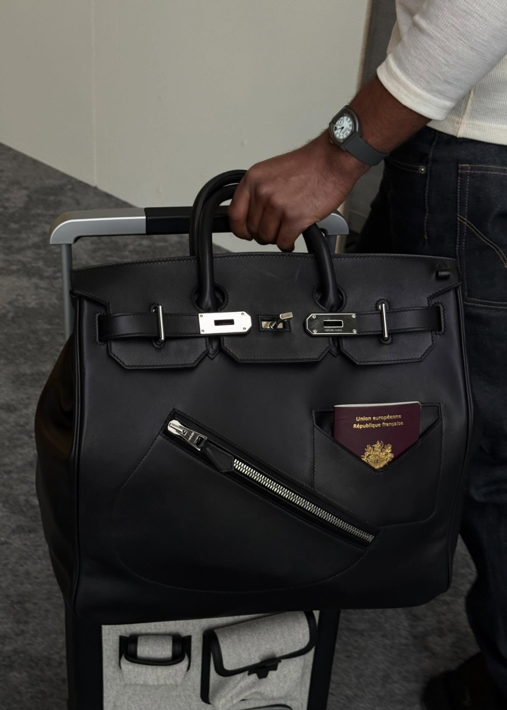 Jules Koundé segura bolsa estruturada de couro preto com zíper e passaporte à mostra, em visual sóbrio e elegante.