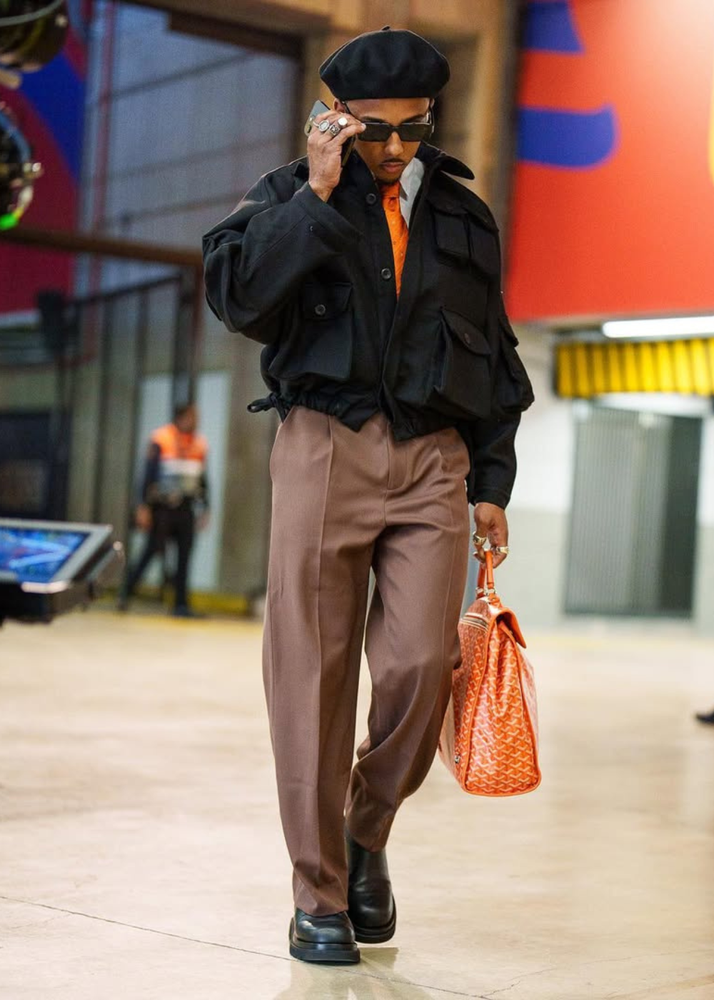 Jules Koundé caminha com boina, jaqueta utilitária preta, calça ampla marrom e bolsa laranja, em visual sofisticado e seguro