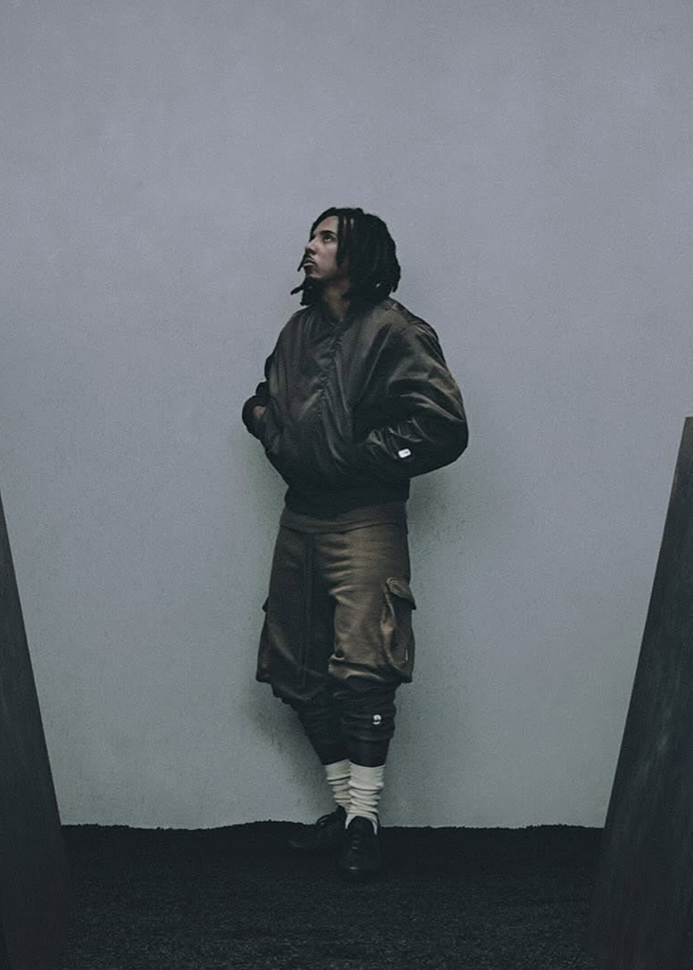 Jules Koundé posa de perfil com bomber acetinada e bermuda cargo marrom, em cenário cinza de clima introspectivo.