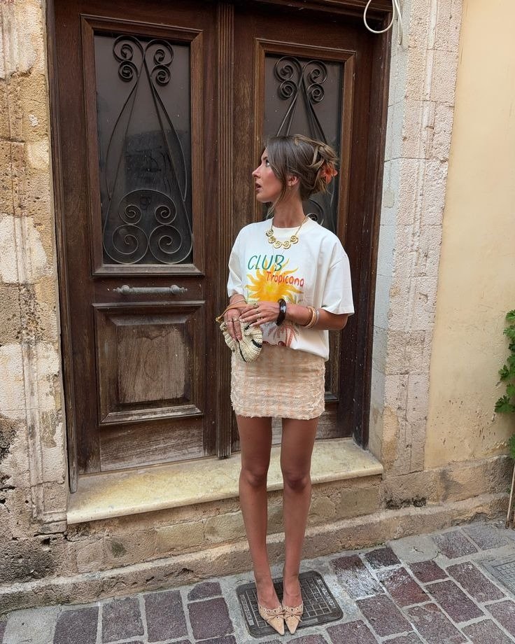 Looks com scarpin bege e minissaia texturizada; modelo posa elegante diante de porta rústica. Texto: CLUB Tropicana