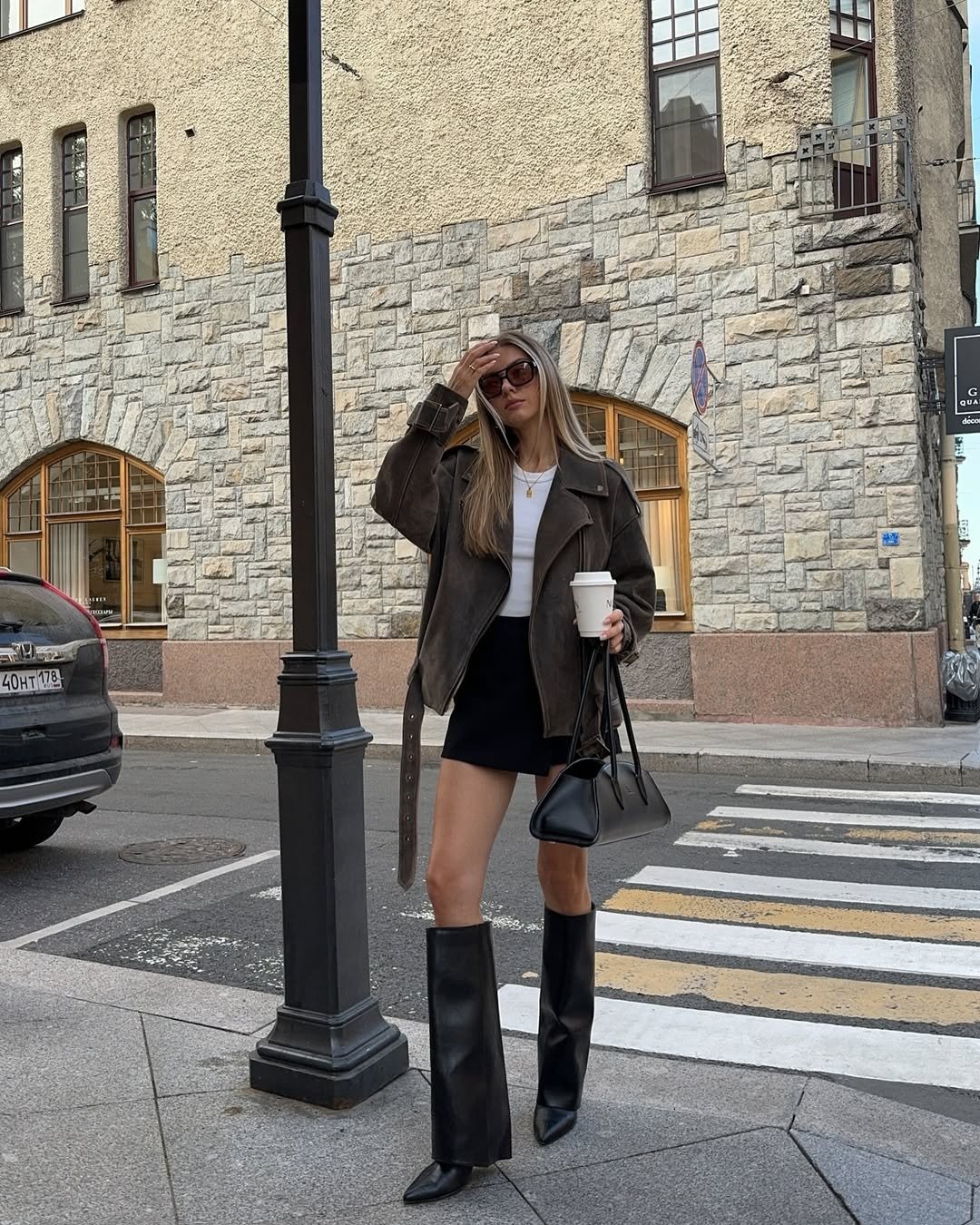 Modelo posa na rua com bota capa preta, minissaia, regata branca e jaqueta marrom oversized em clima urbano sofisticado