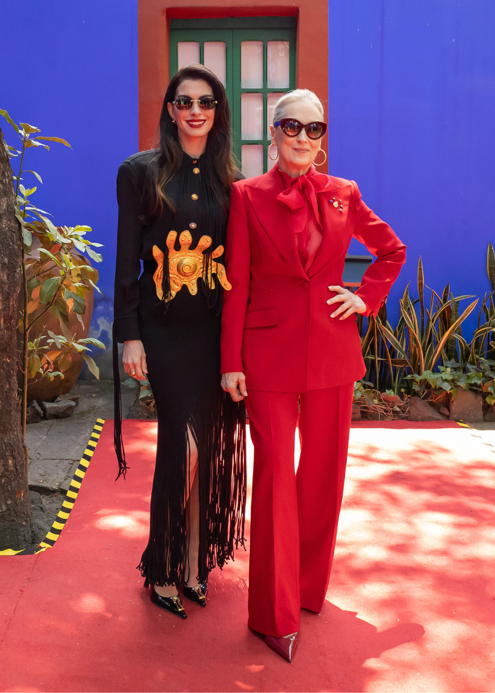 Method dressing em dupla: mulher posa de vestido preto com franjas e outra de alfaiataria vermelha, clima sofisticado.