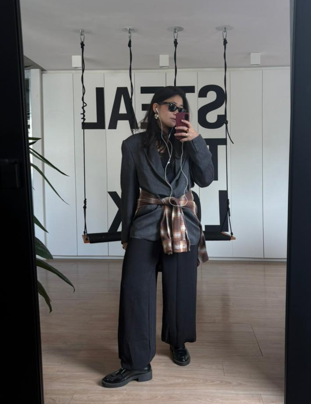 Looks de escritório com blazer cinza, calça ampla preta e camisa xadrez na cintura; modelo posa com elegância urbana.