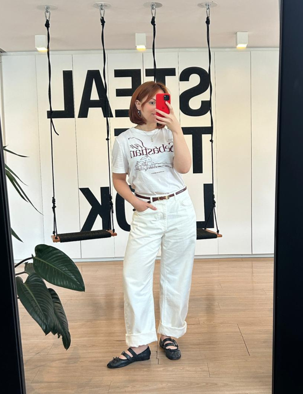 Looks de escritório casuais com camiseta e calça reta brancas, cinto marrom e sapatilha preta; modelo posa leve no espelho.