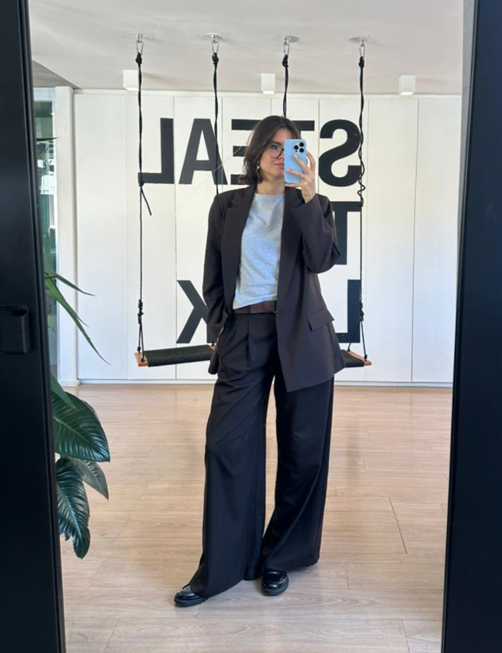 Looks de escritório com blazer e calça ampla pretos, camiseta cinza; modelo posa ao espelho com elegância urbana.