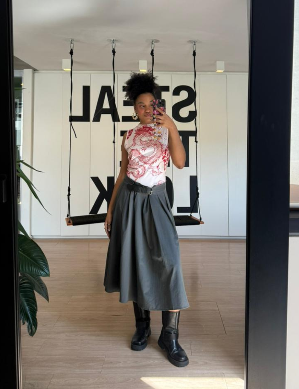 Looks de escritório com blusa estampada, saia midi cinza ampla e bota preta; modelo posa em selfie elegante.