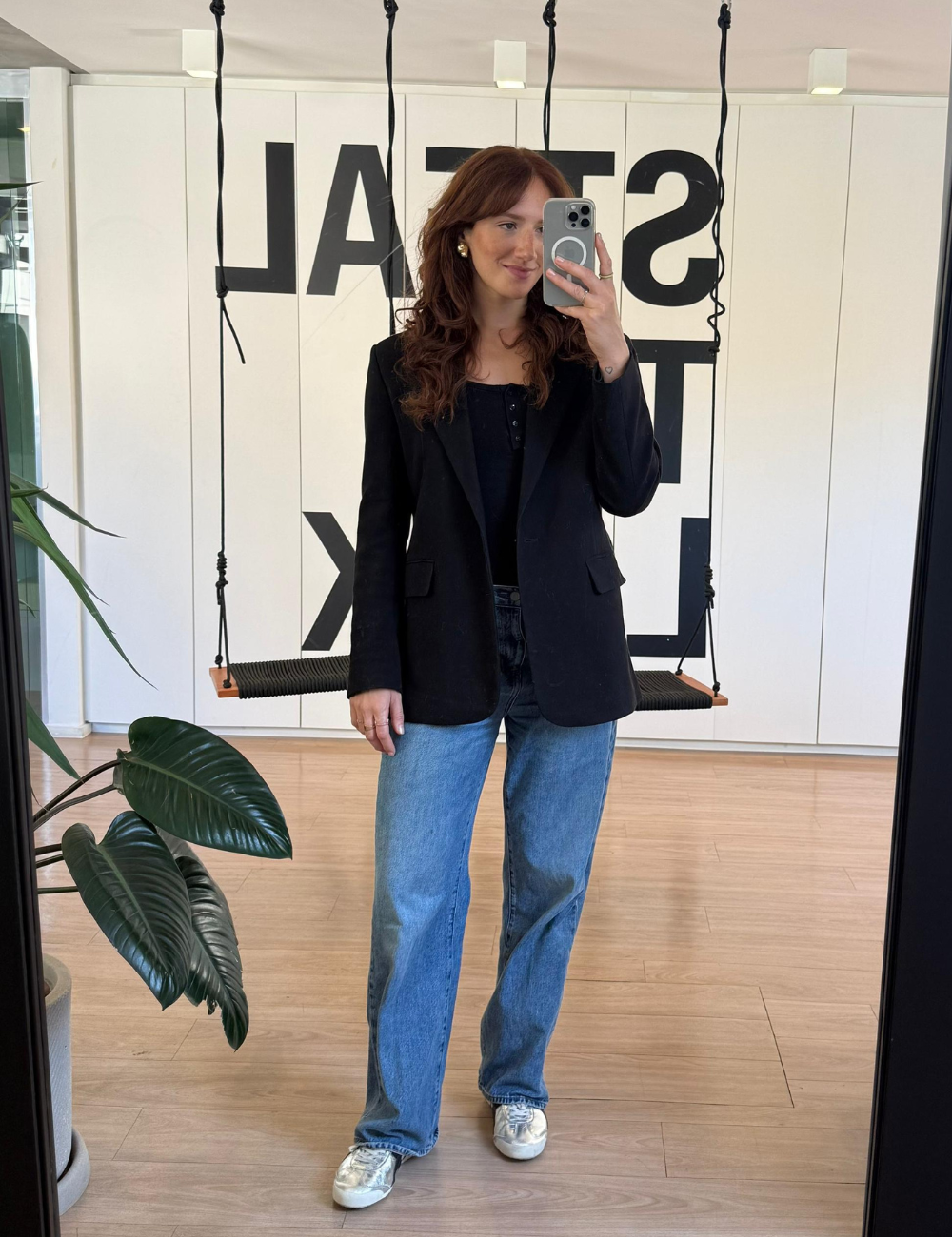 Looks de escritório com blazer preto, jeans reto azul e tênis prata; modelo posa ao espelho em clima casual elegante.