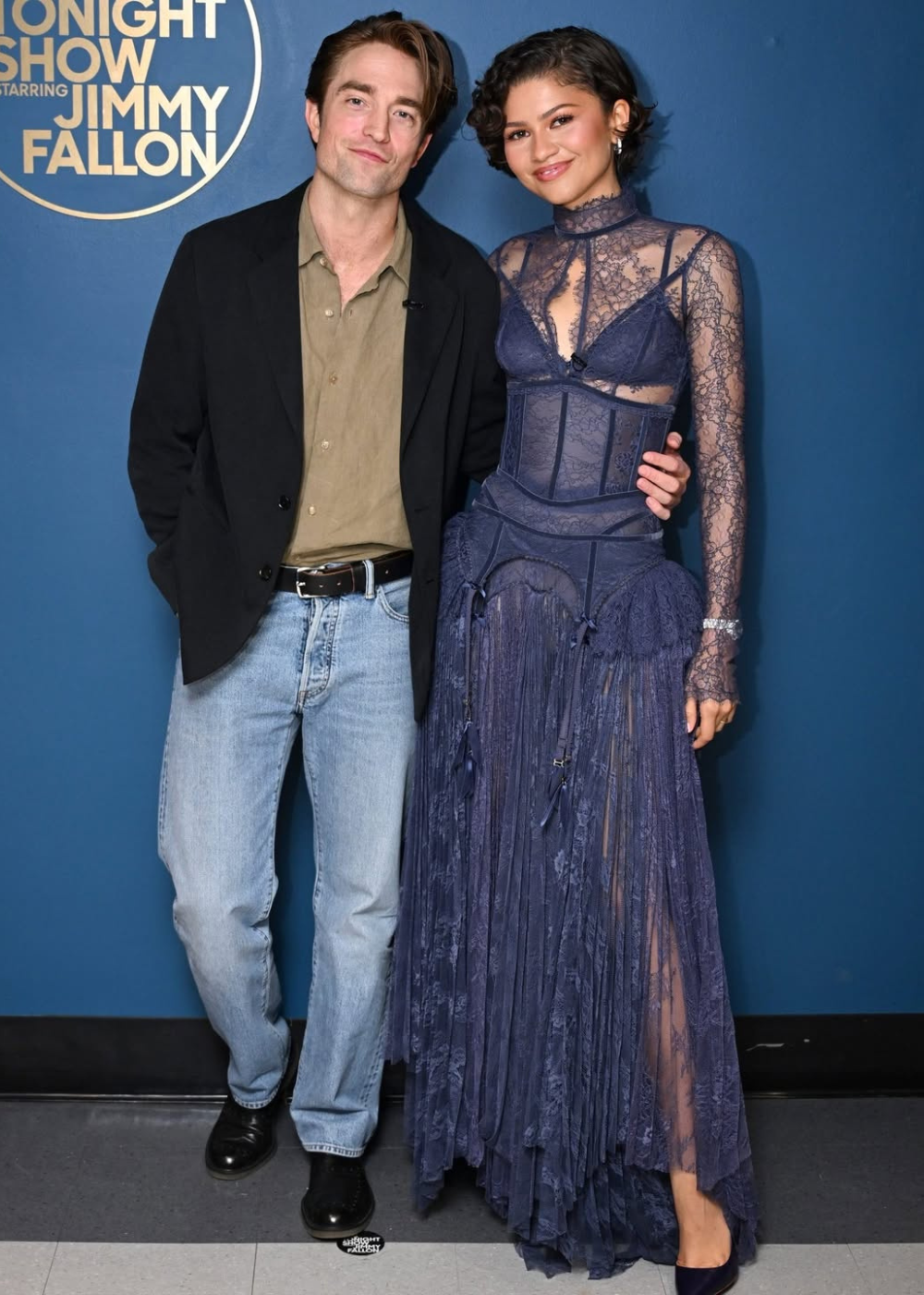 Casal posa em method dressing; ela veste renda azul marinho com corset e franjas, ele usa blazer preto e jeans.