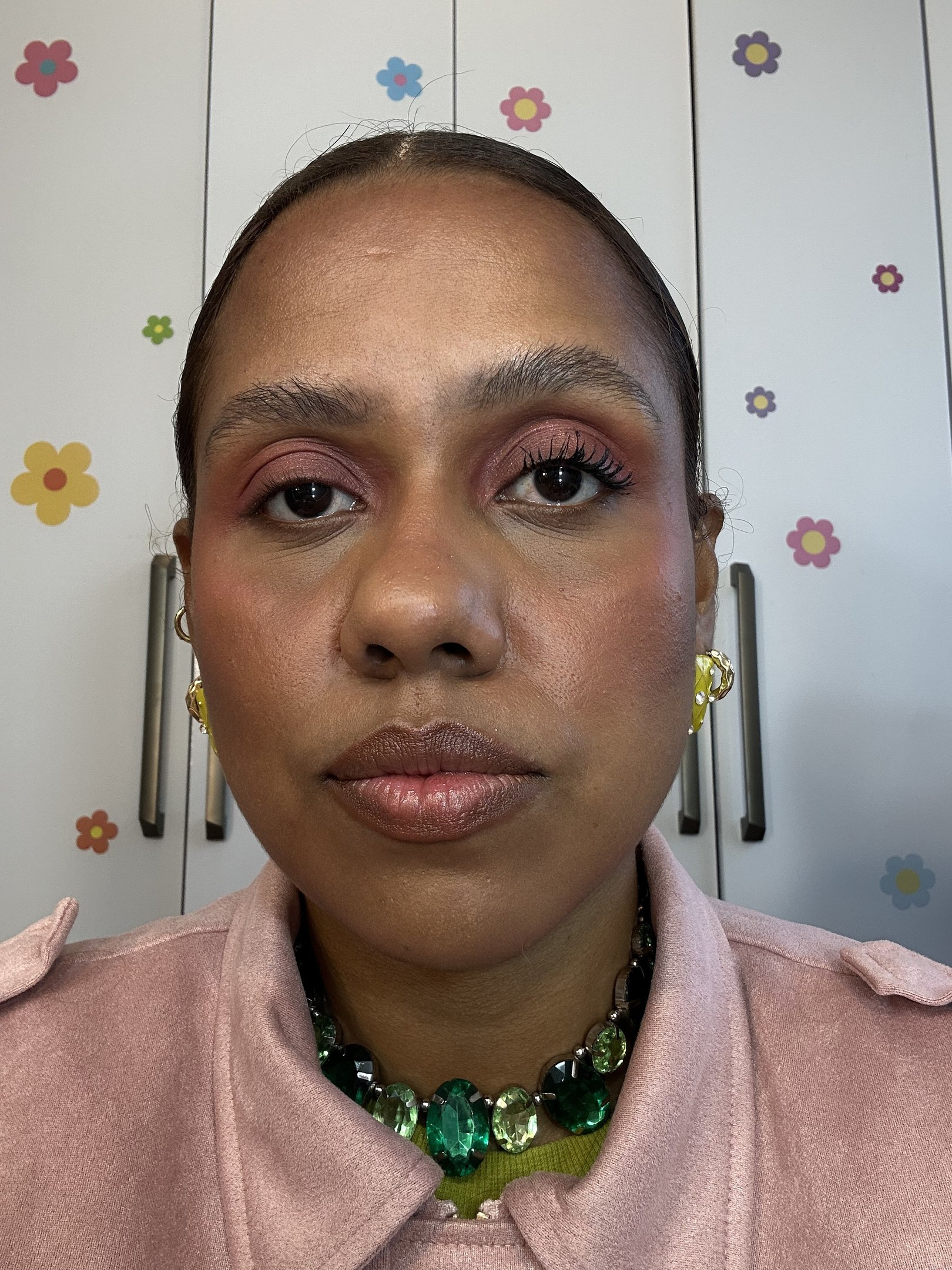 Máscara de cílios da Essence destaca olhar com volume; modelo posa de frente com sombra rosa, jaqueta rosé e colar verde.