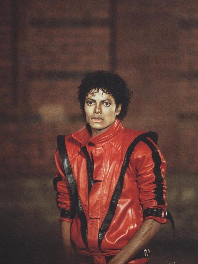 Michael Jackson posa com jaqueta de couro vermelha e preta de ombros marcados, em clima icônico e intenso.