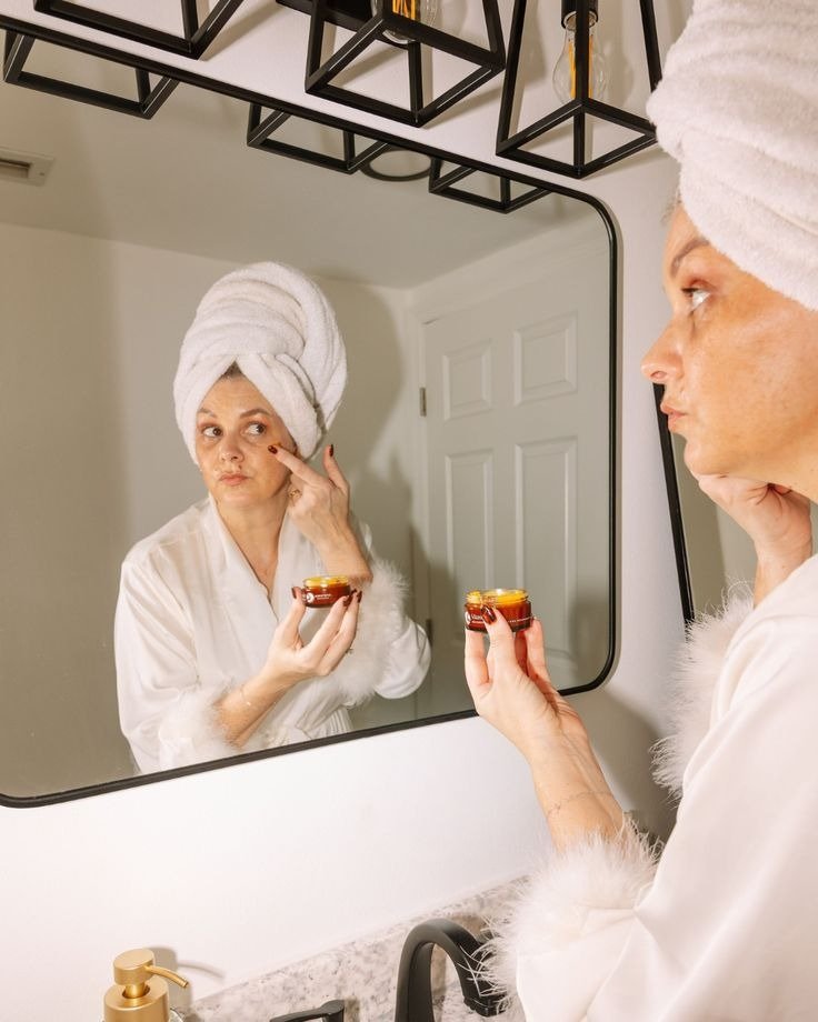 Melhor sérum para pele madura em cena de autocuidado: mulher de roupão branco aplica creme diante do espelho, clima sereno.