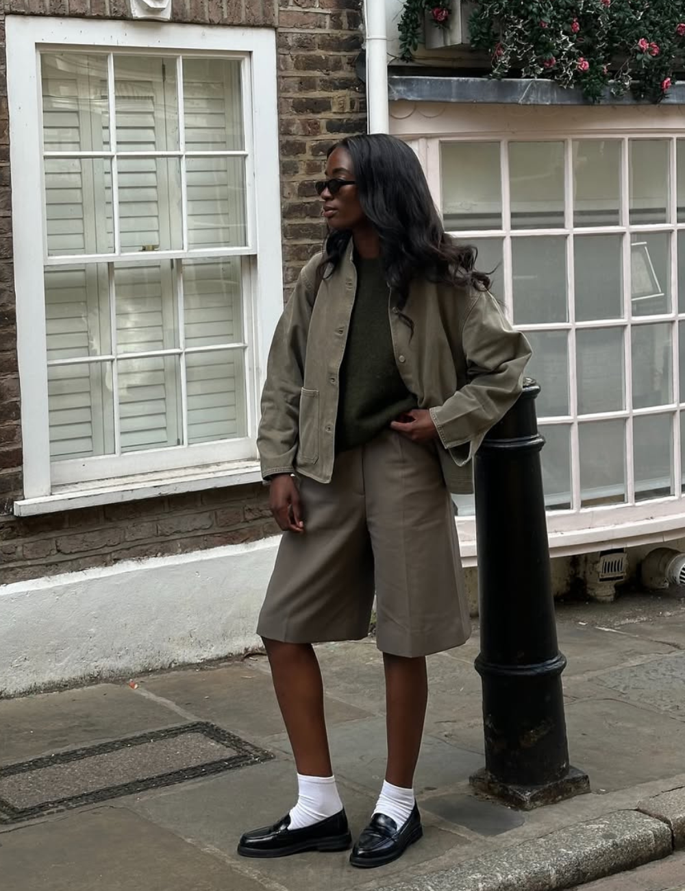 Modelo posa na calçada com mocassins femininos estilosos pretos, meias brancas e alfaiataria cáqui em clima urbano elegante.