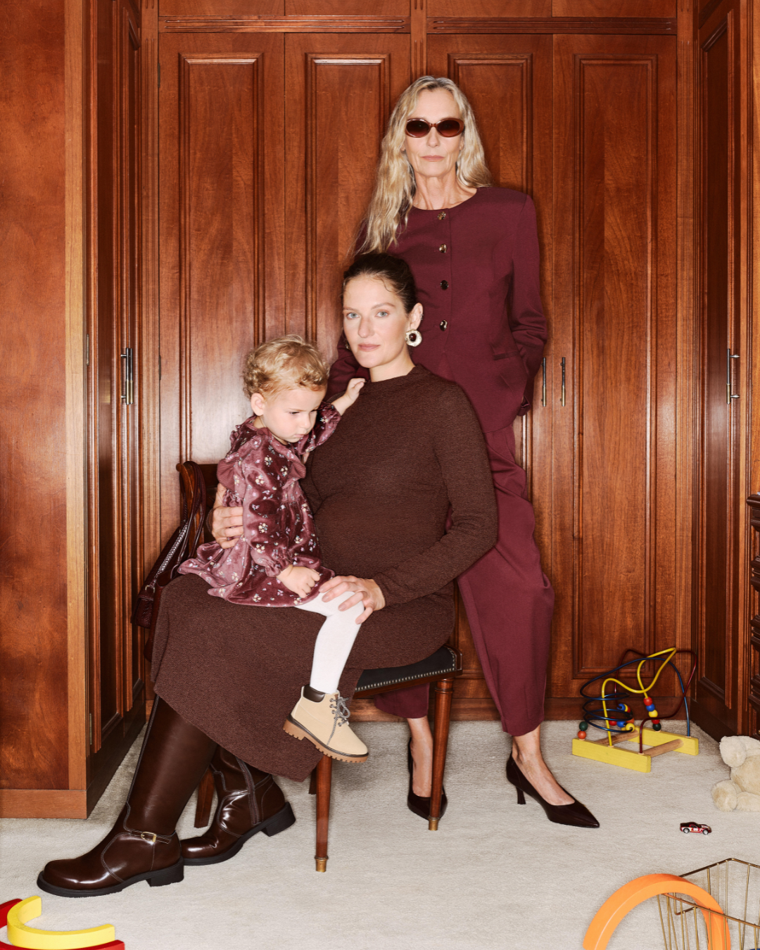 Moda e maternidade em tons vinho e marrom: mãe grávida senta com criança no colo, enquanto mulher posa ao fundo.