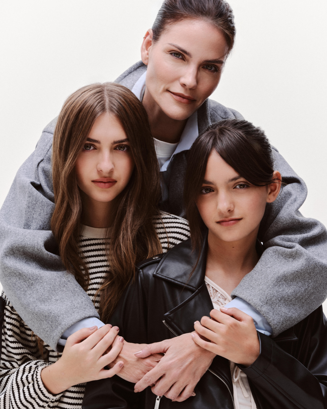 Moda e maternidade em retrato afetuoso: mulher posa com jovens em cinza, listras e couro preto sobre fundo neutro.