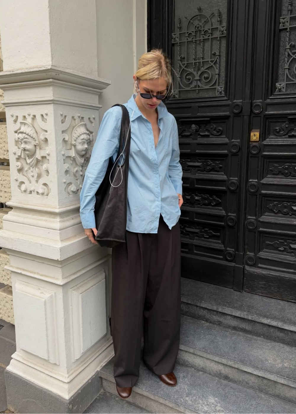 Looks com camisa azul feminina: modelo posa com camisa ampla, calça marrom fluida e bolsa preta em escadaria elegante.