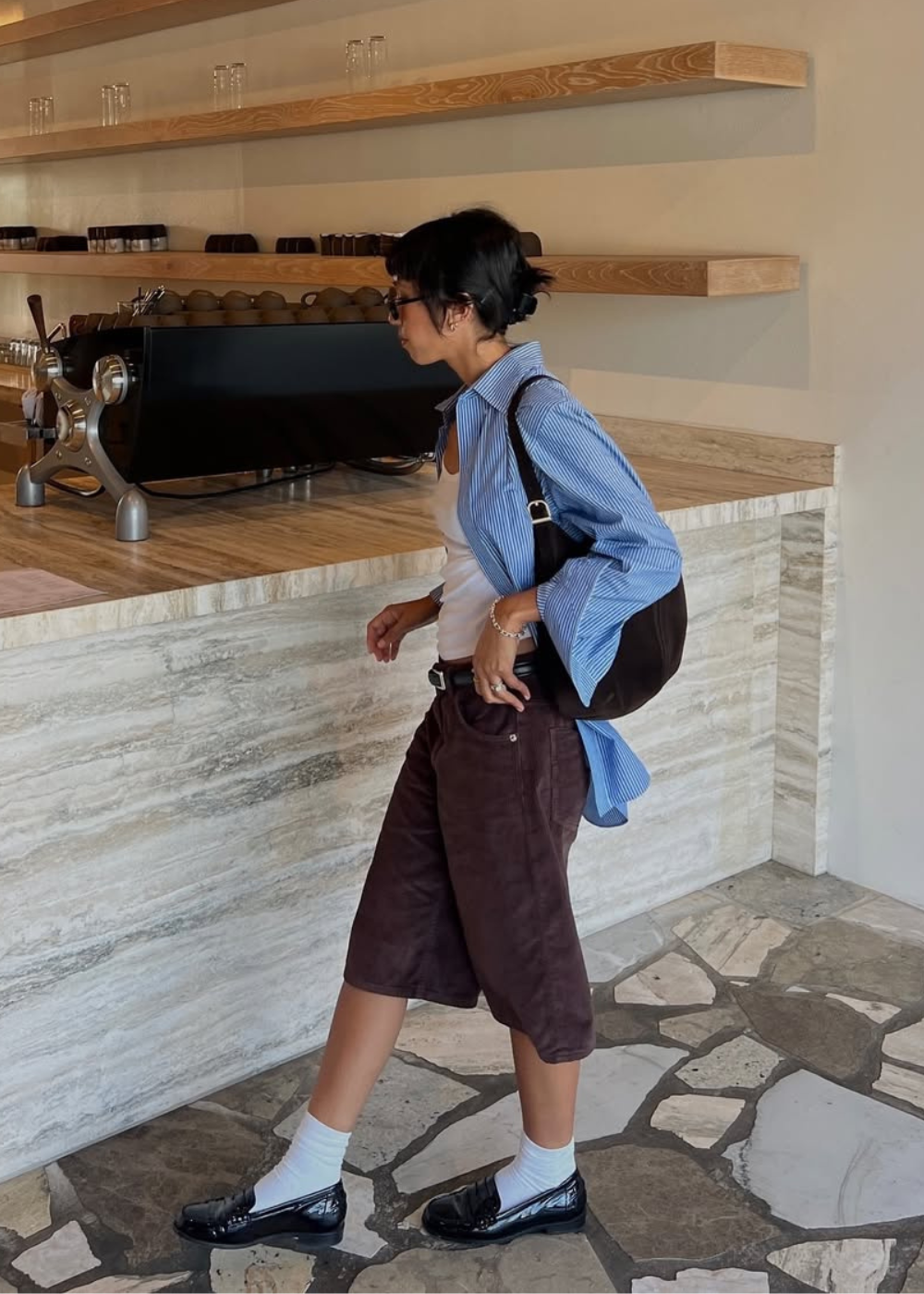 Looks com camisa azul feminina: modelo posa em café com listras soltas, regata branca, bermuda marrom e mood casual elegante