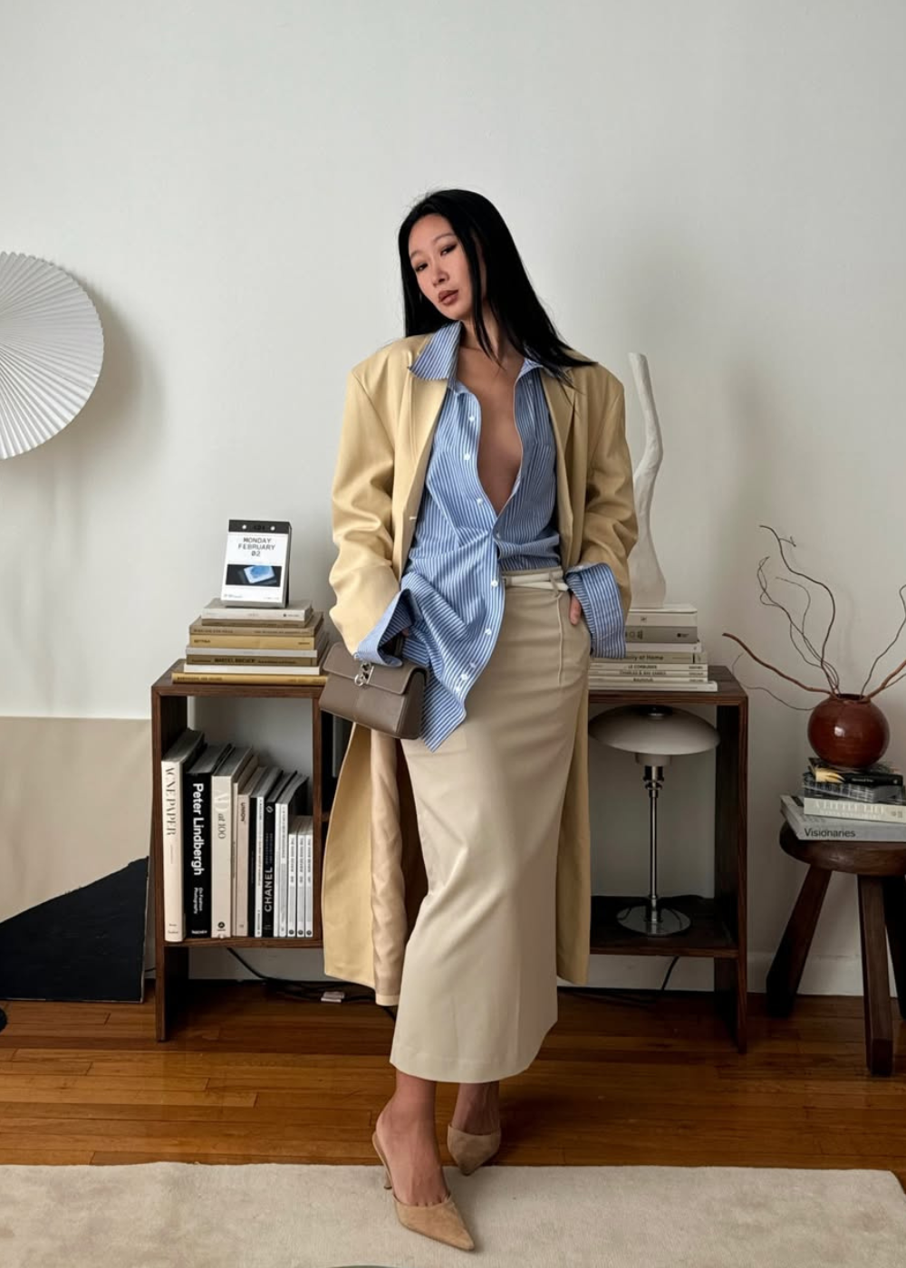 Looks com camisa azul feminina: modelo posa com trench bege, saia midi acetinada e scarpin em sala minimalista elegante.