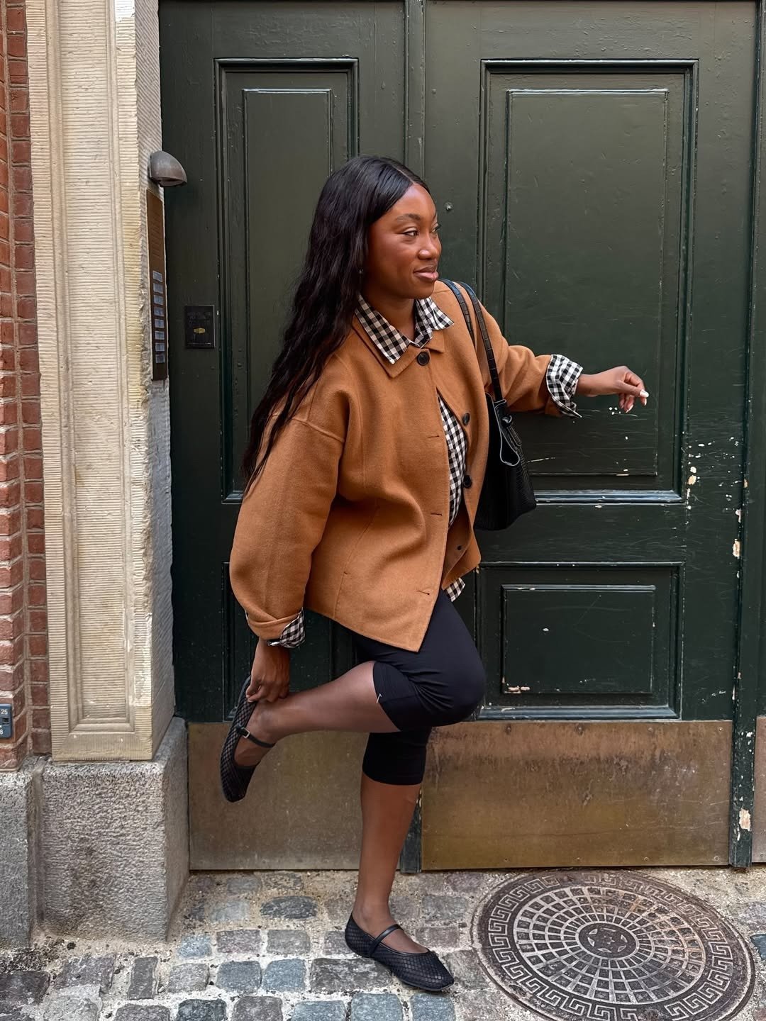 Modelo posa com sapatilha de tela preta, casaco caramelo e xadrez vichy diante de porta verde, em clima casual elegante.