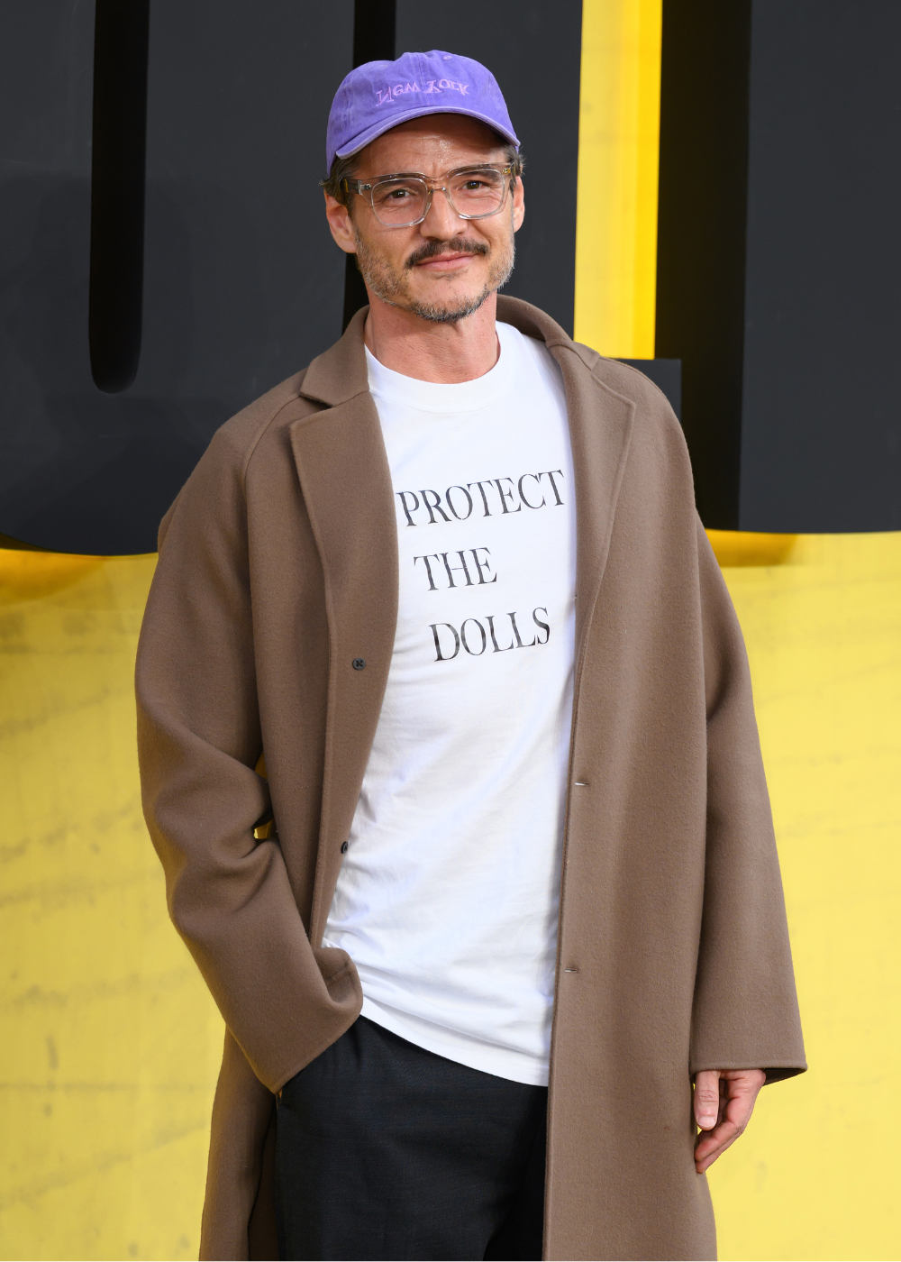 Modelo posa com camiseta branca de algodão, casaco marrom e boné lilás diante de fundo amarelo.