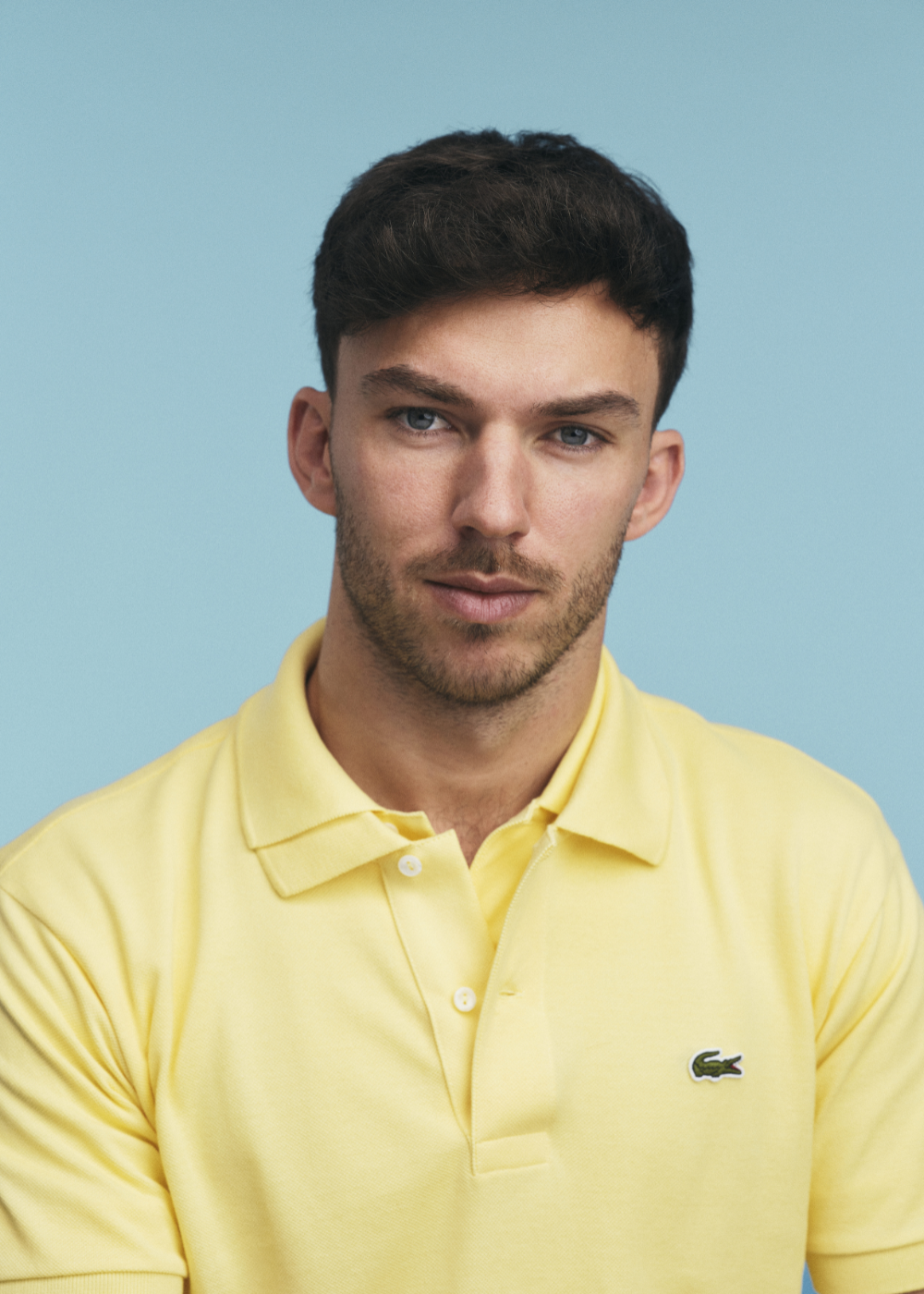 Pierre Gasly posa em polo amarela de piquê com corte clássico, sobre fundo azul-claro, em clima sereno e confiante.
