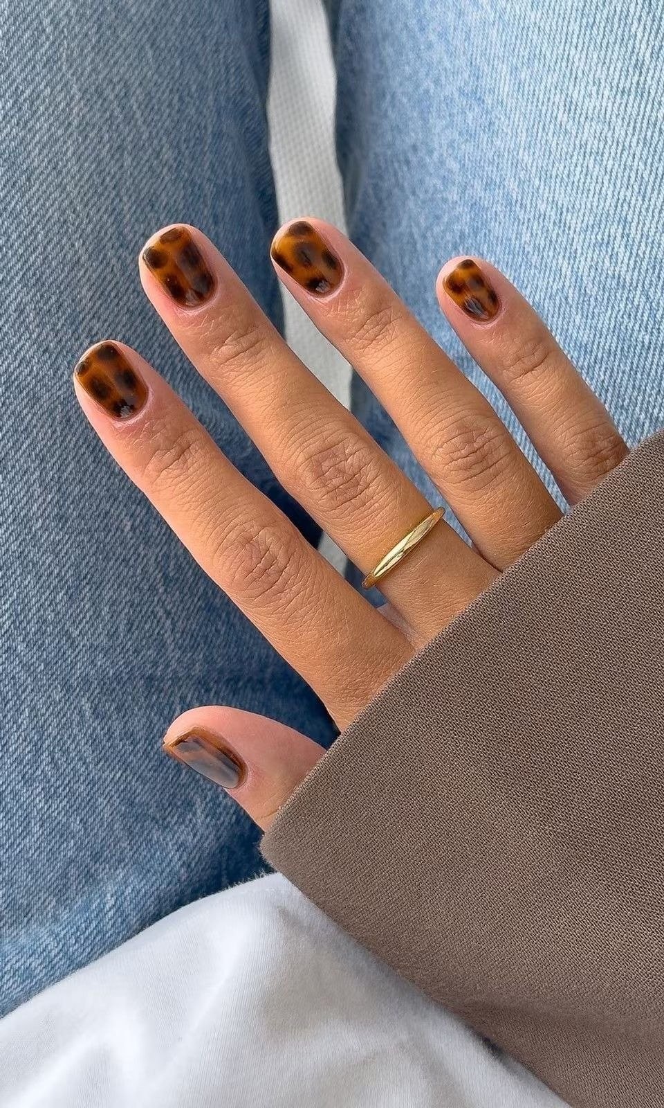 Unhas mais populares em estampa tartaruga marrom, mão posa com anel dourado sobre jeans e manga bege, clima elegante.