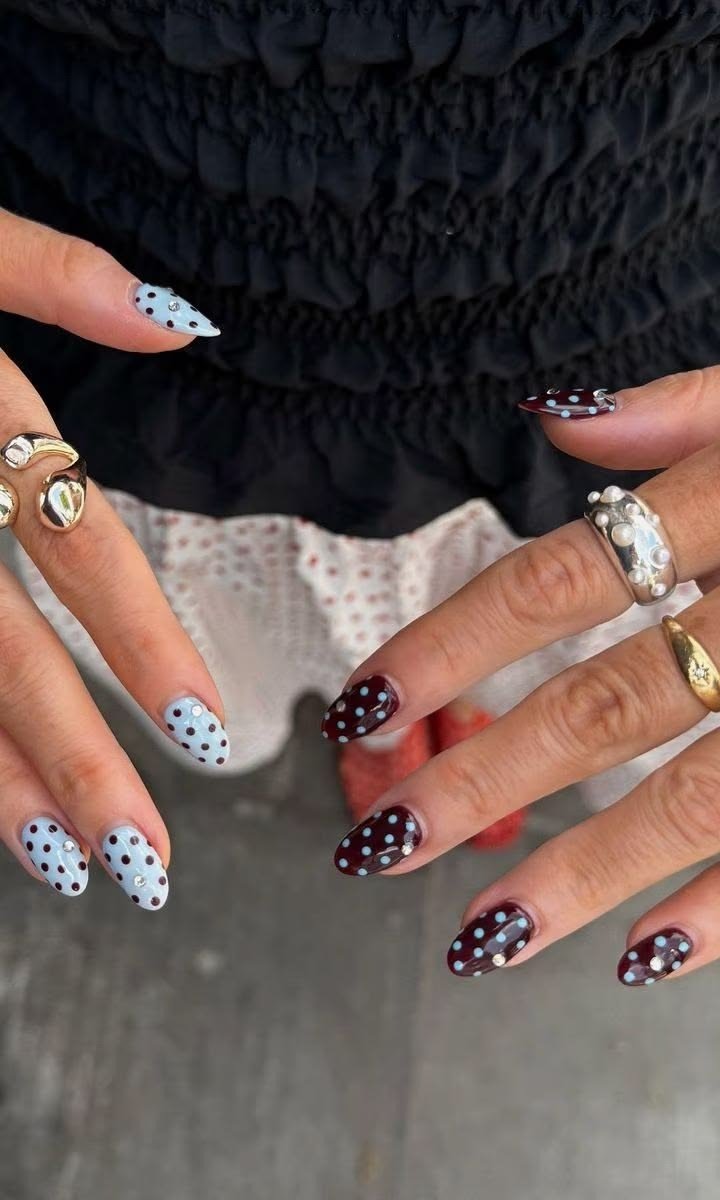 Unhas mais populares em formato amendoado com poás azul-claro e vinho, anéis metálicos e blusa preta criam estilo moderno.