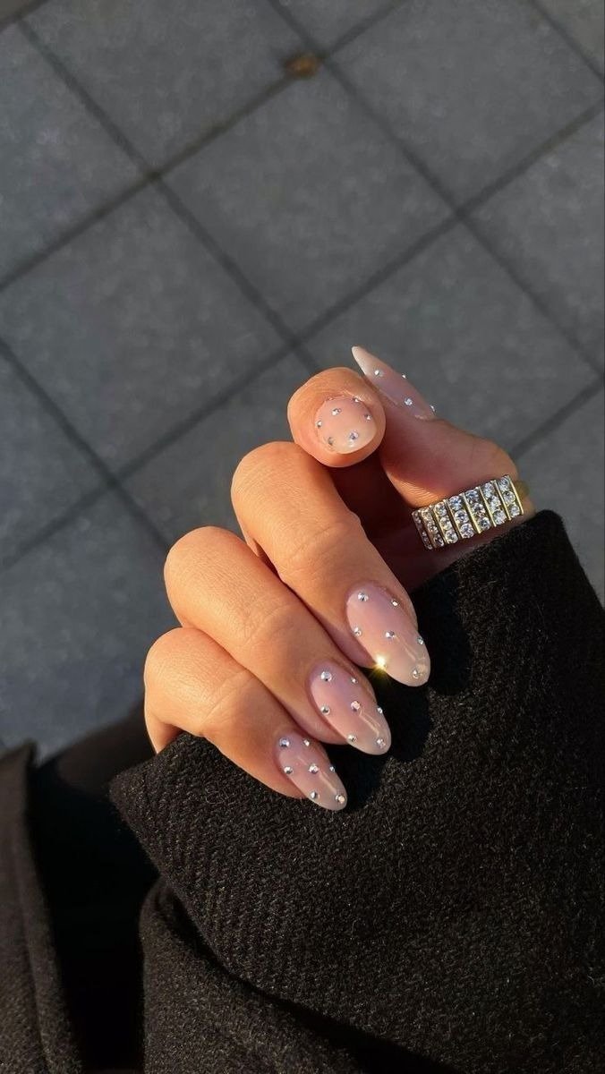 Unhas mais populares em nude brilhante com strass, mão posa com tricô preto e anel dourado sob luz elegante.