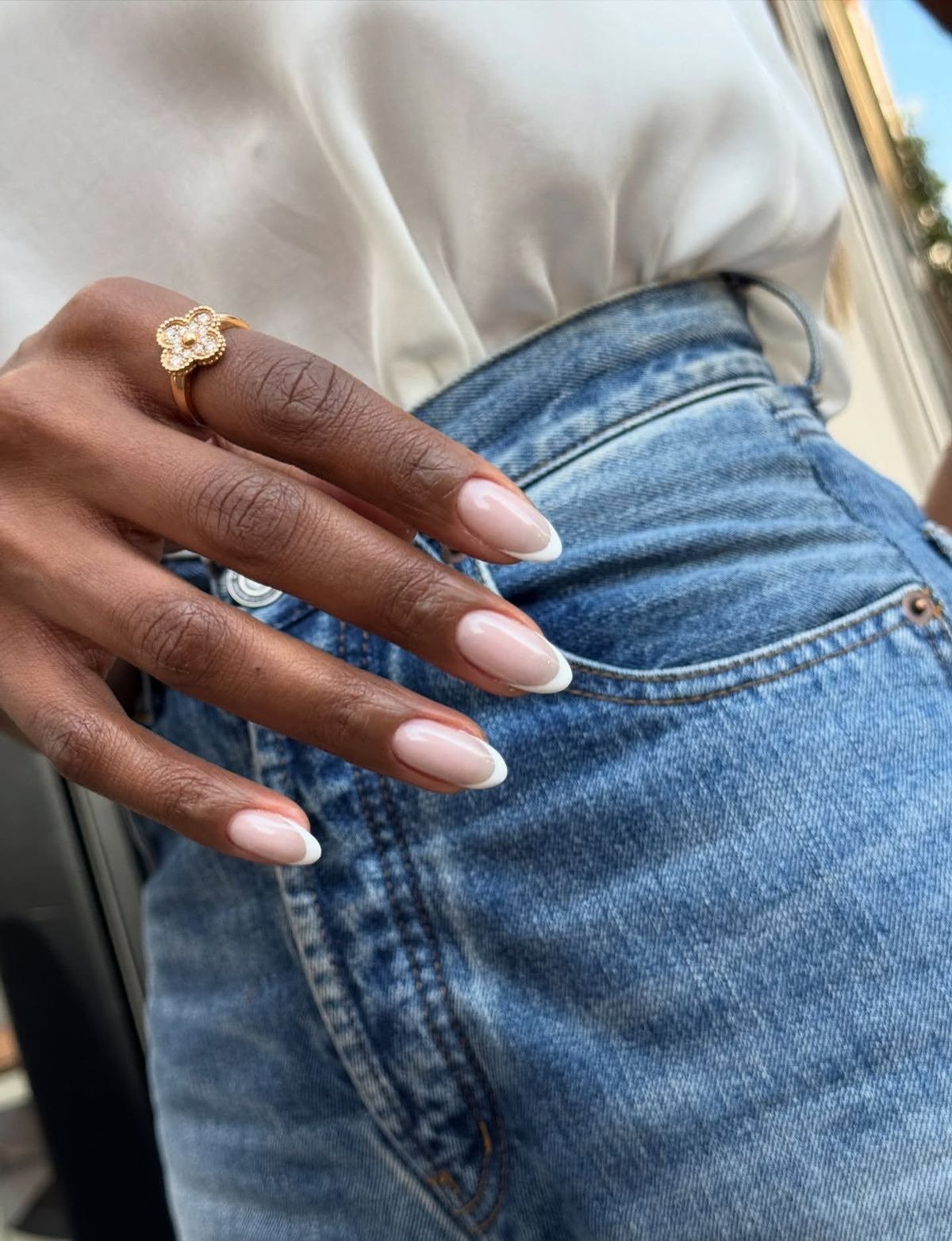 Unhas mais populares em francesinha branca oval brilham na mão com anel floral dourado, jeans azul e blusa creme elegante