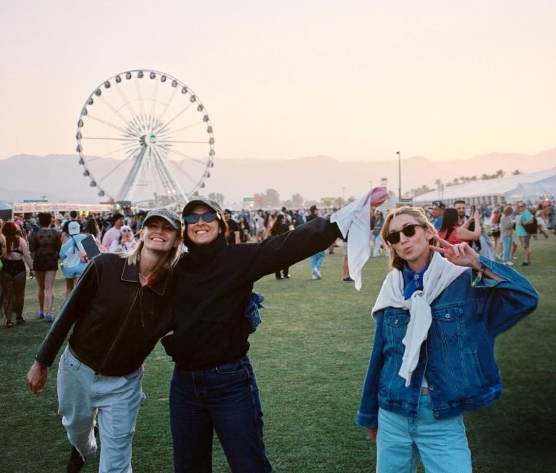 Amigas posam sorrindo em festival ao pôr do sol, com jaquetas escuras e jeans azul, clima leve diante da roda-gigante