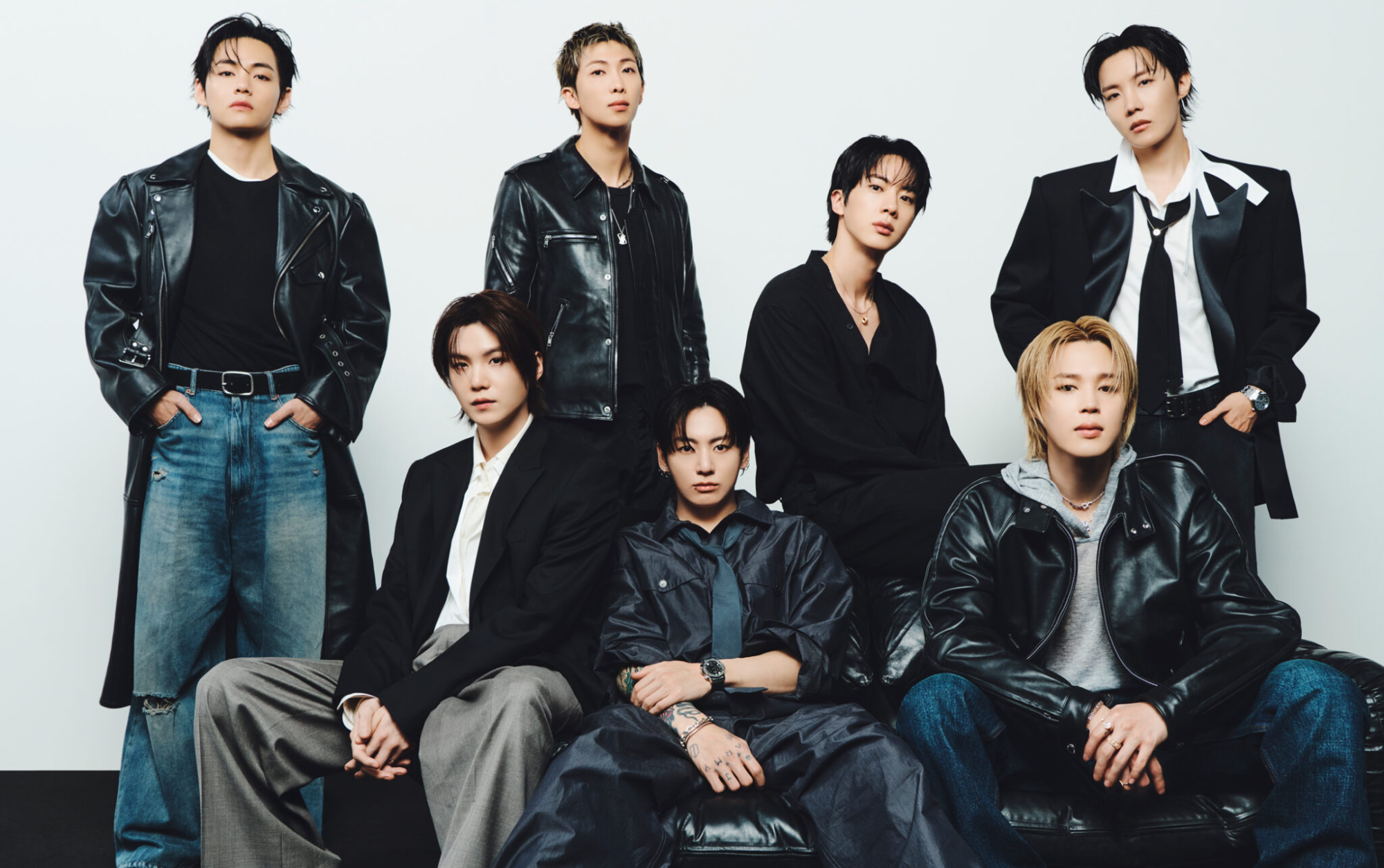 BTS posa em grupo com couro preto, alfaiataria e jeans largos em fundo claro, criando clima elegante e intenso.