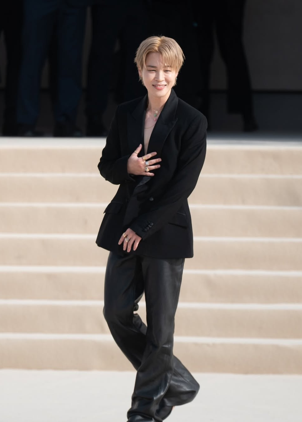 Jimin, do BTS, posa sorrindo em blazer preto de alfaiataria e calça ampla de couro, com joias douradas em escadaria clara.