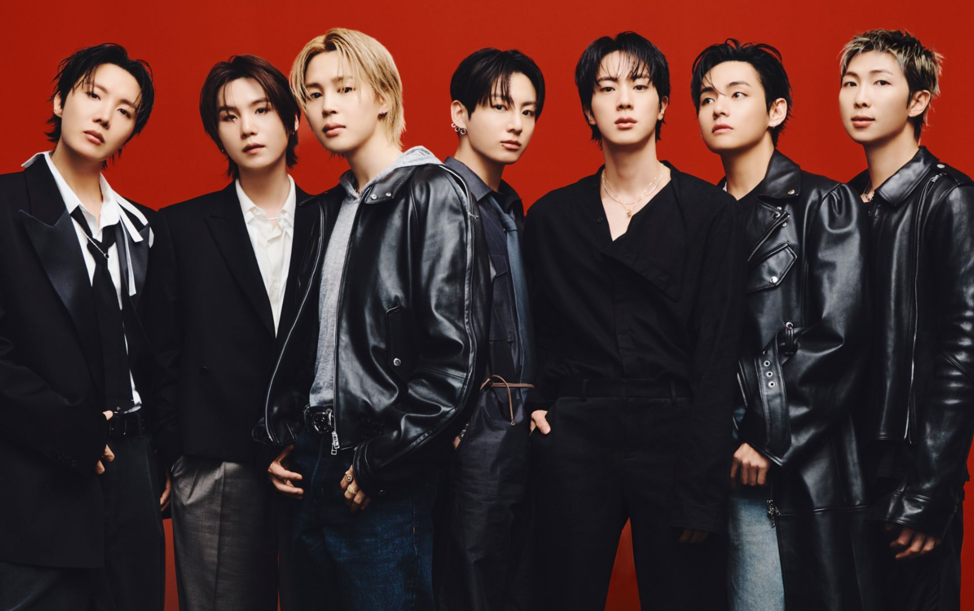 BTS posa em grupo com alfaiataria preta e jaquetas de couro diante de fundo vermelho, em clima marcante e confiante.