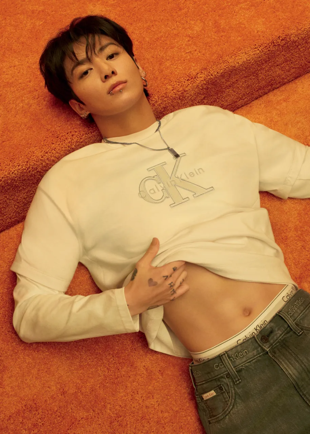 BTS posa deitado em tapete laranja com moletom branco, jeans e cintura Calvin Klein à mostra; clima sensual.