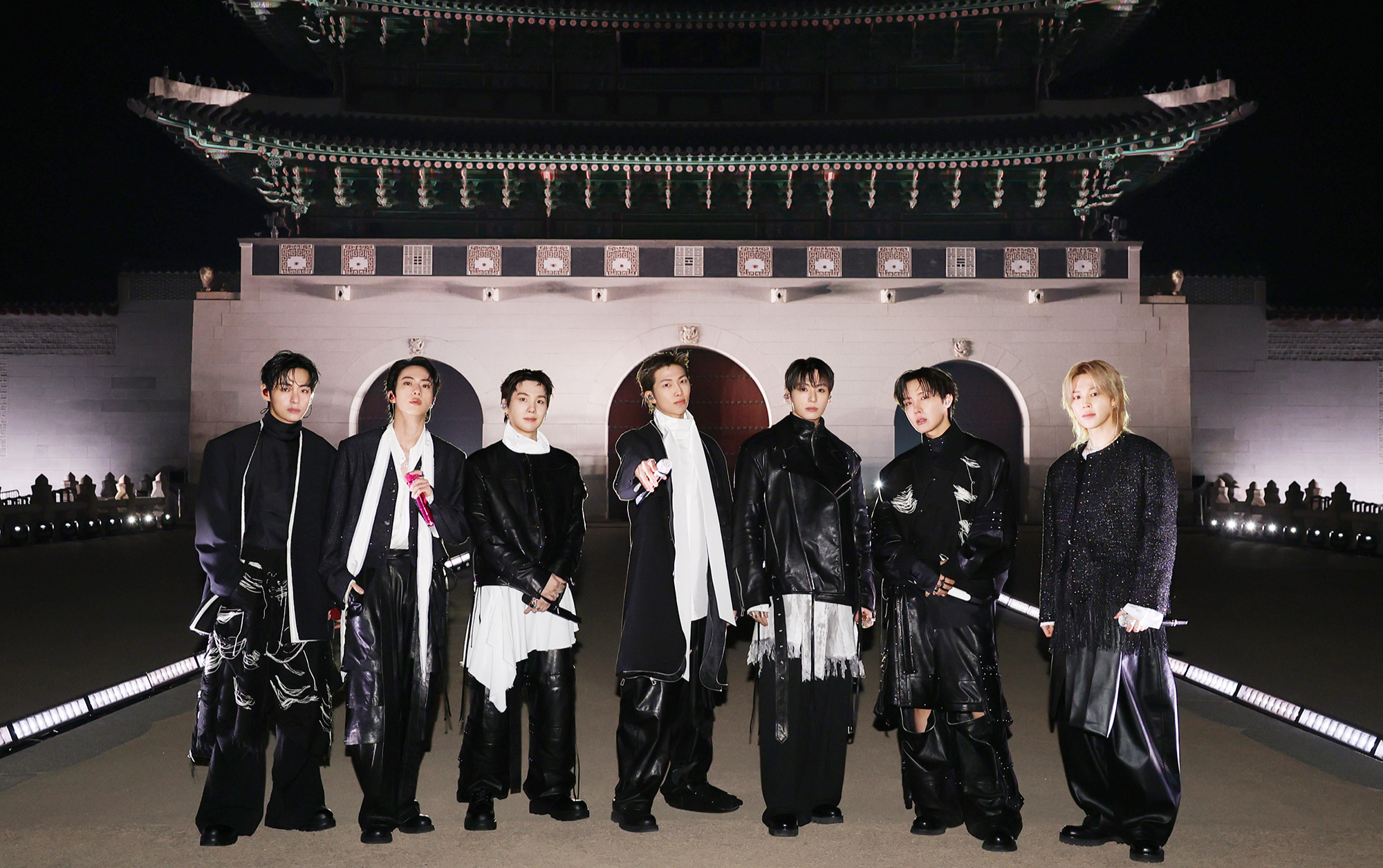 BTS posa em grupo com looks pretos de couro e camadas fluidas diante de palácio iluminado, em clima solene e poderoso