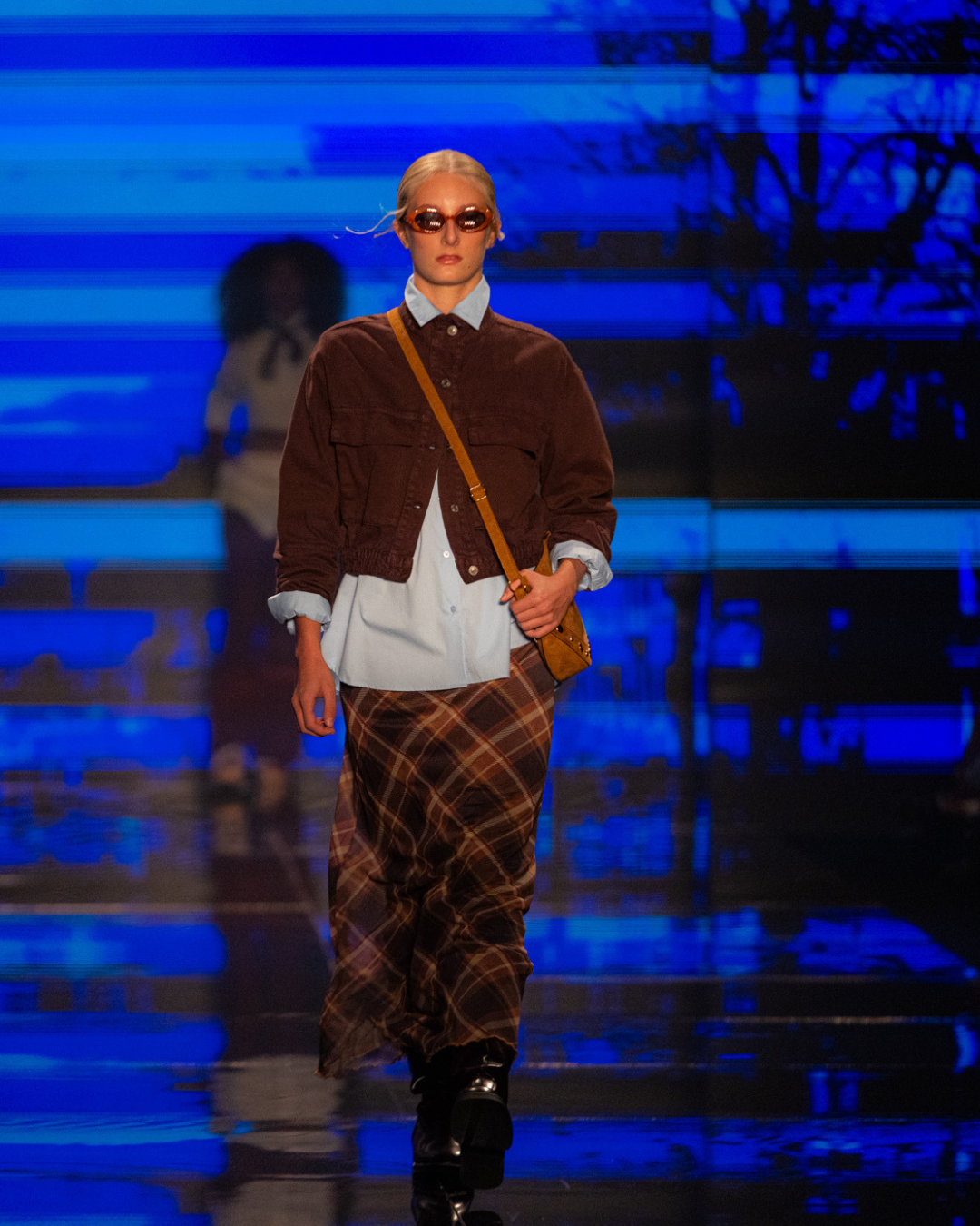 Modelo posa na passarela com jaqueta marrom, camisa azul e saia xadrez, cores e estampas tendência do outono inverno 2026.