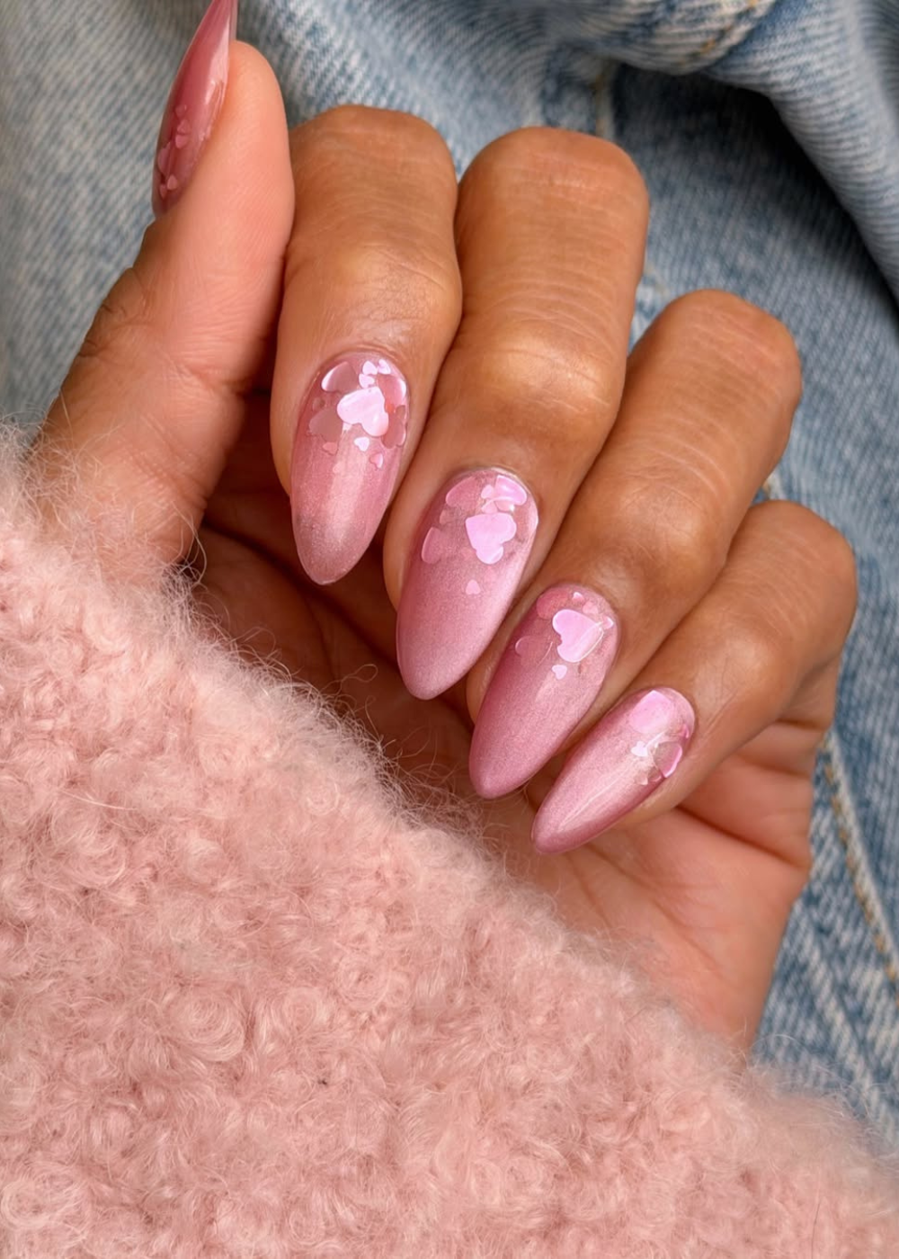 Rosa pastel em unhas amendoadas peroladas com corações, mão posa sobre tricô felpudo e jeans em clima delicado.