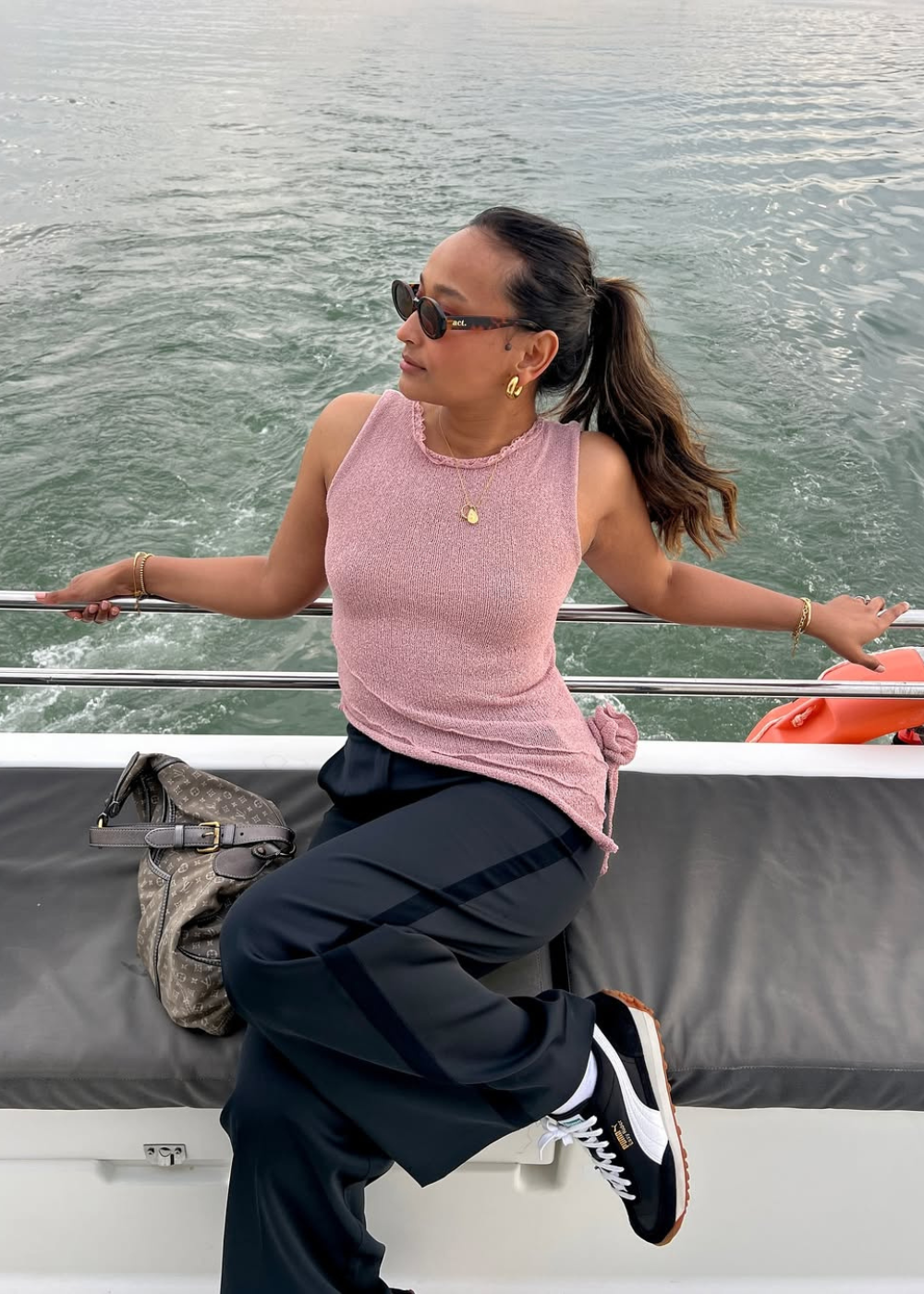 Rosa pastel em regata de tricô; modelo posa no barco com calça ampla preta, tênis e clima leve à beira d’água