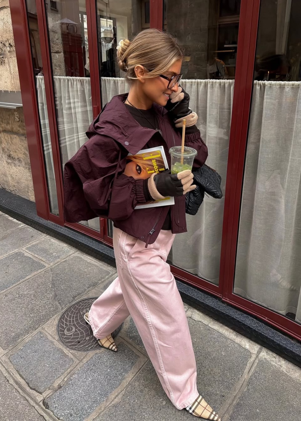 Modelo posa caminhando com calça rosa pastel acetinada, jaqueta vinho e luvas, em clima urbano elegante.